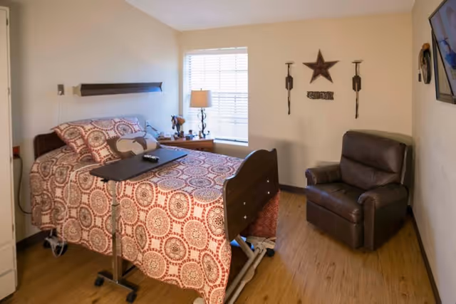 A small bedroom in a senior living facility with a hospital-style bed covered in a patterned red and white bedspread. Next to the bed is a wooden nightstand with a lamp and decorative items. A brown leather recliner chair is positioned against the wall, which is decorated with a star and two candle holders. A flat-screen TV is mounted on the opposite wall. The room has wood flooring and a window with blinds letting in natural light.