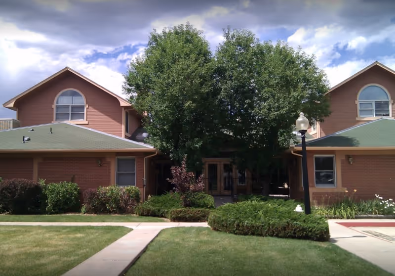 Exterior view of a two-story brick building with green roofs, surrounded by well-maintained bushes and trees. A concrete pathway leads to the entrance, which is partially obscured by large trees. The sky is partly cloudy.