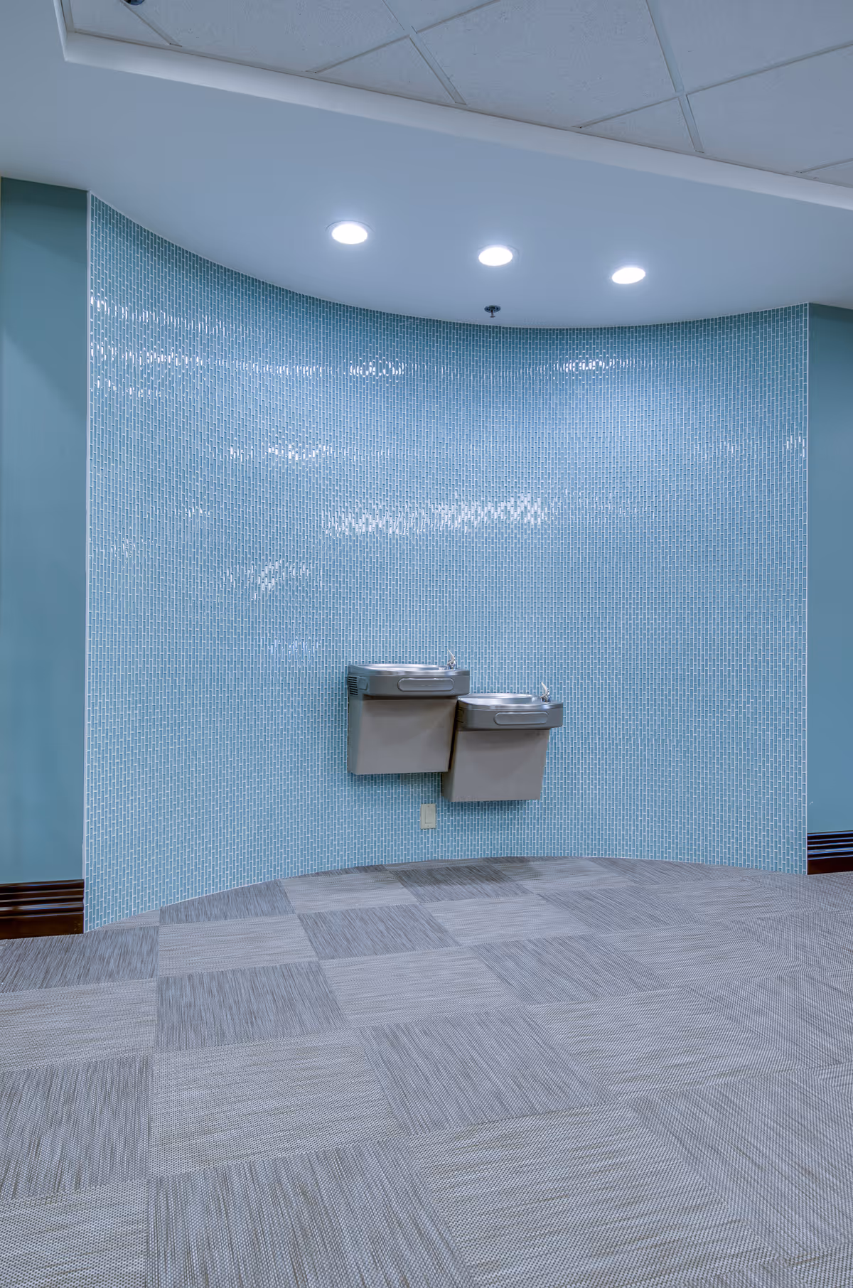 Two wall-mounted drinking fountains of different heights against a curved light blue tiled wall in an indoor space with a patterned carpet floor and recessed ceiling lights.