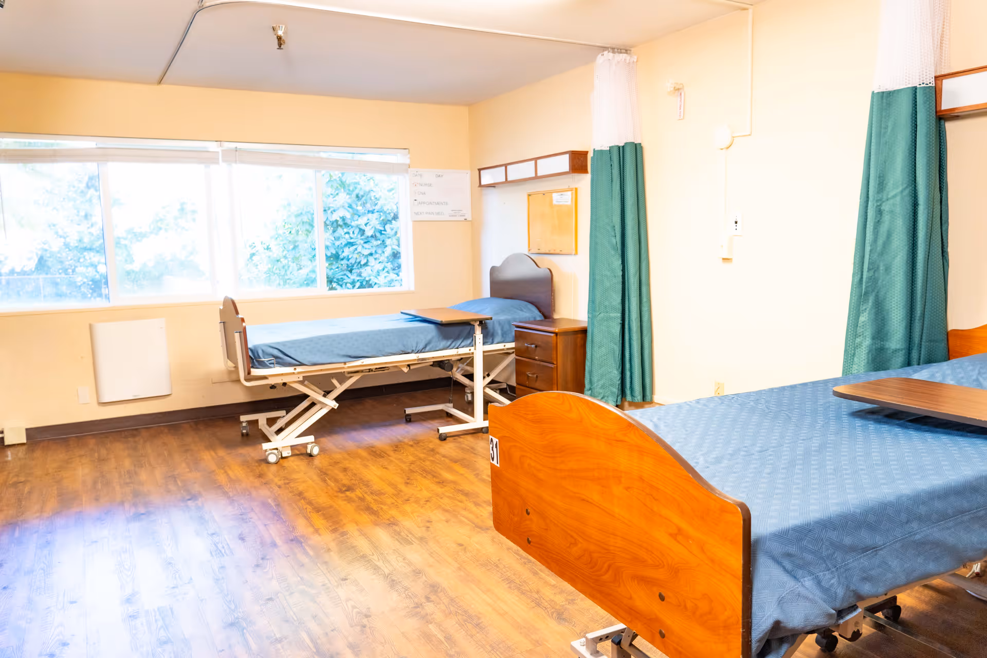Bright shared patient room with two hospital beds, bedside furniture, privacy curtains, and large windows.