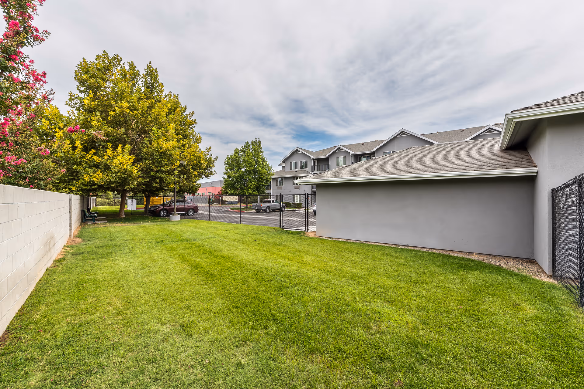 A grassy outdoor area with a few trees and a concrete wall on the left side. There is a building with a gray exterior and a sloped roof on the right side. In the background, there is a parking lot with several cars and a multi-story building under a cloudy sky.