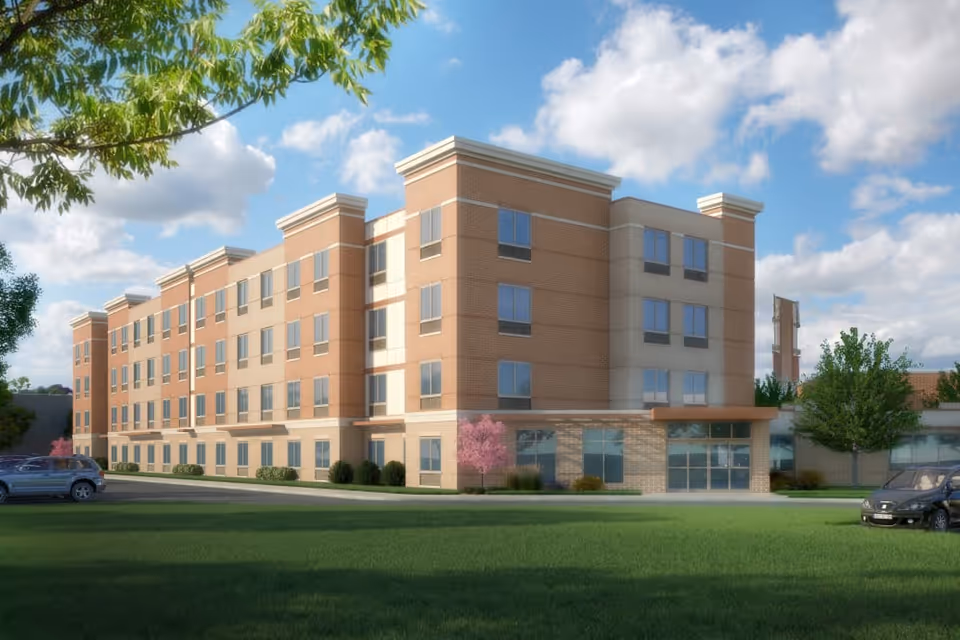 Exterior view of a modern four-story brick building with large windows, surrounded by green grass, trees, and a few parked cars under a partly cloudy sky.
