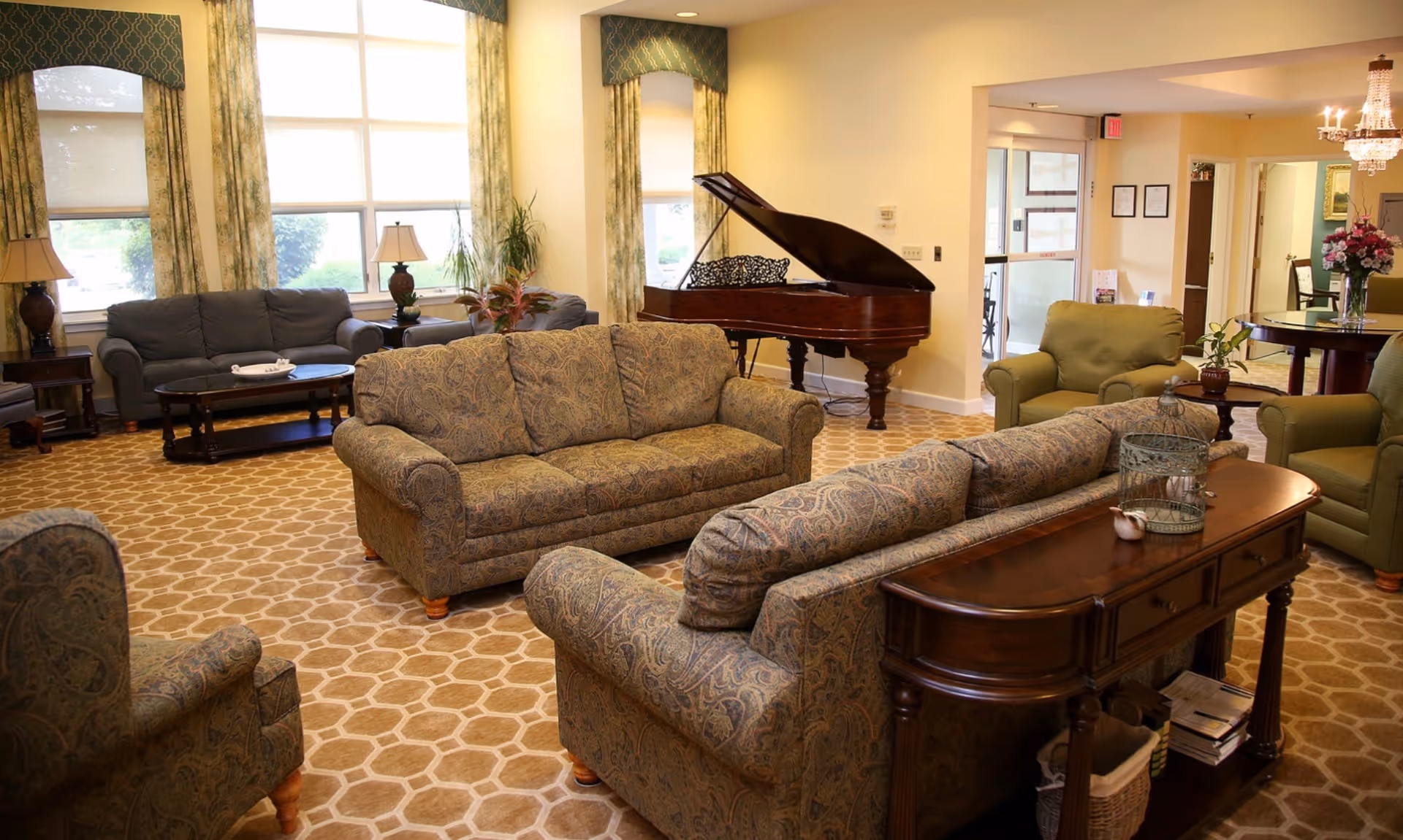 A spacious living room area with patterned sofas and armchairs arranged around wooden tables. Large windows with floral curtains let in natural light. A grand piano is positioned against one wall. The room has a patterned carpet and warm lighting, creating a cozy atmosphere.