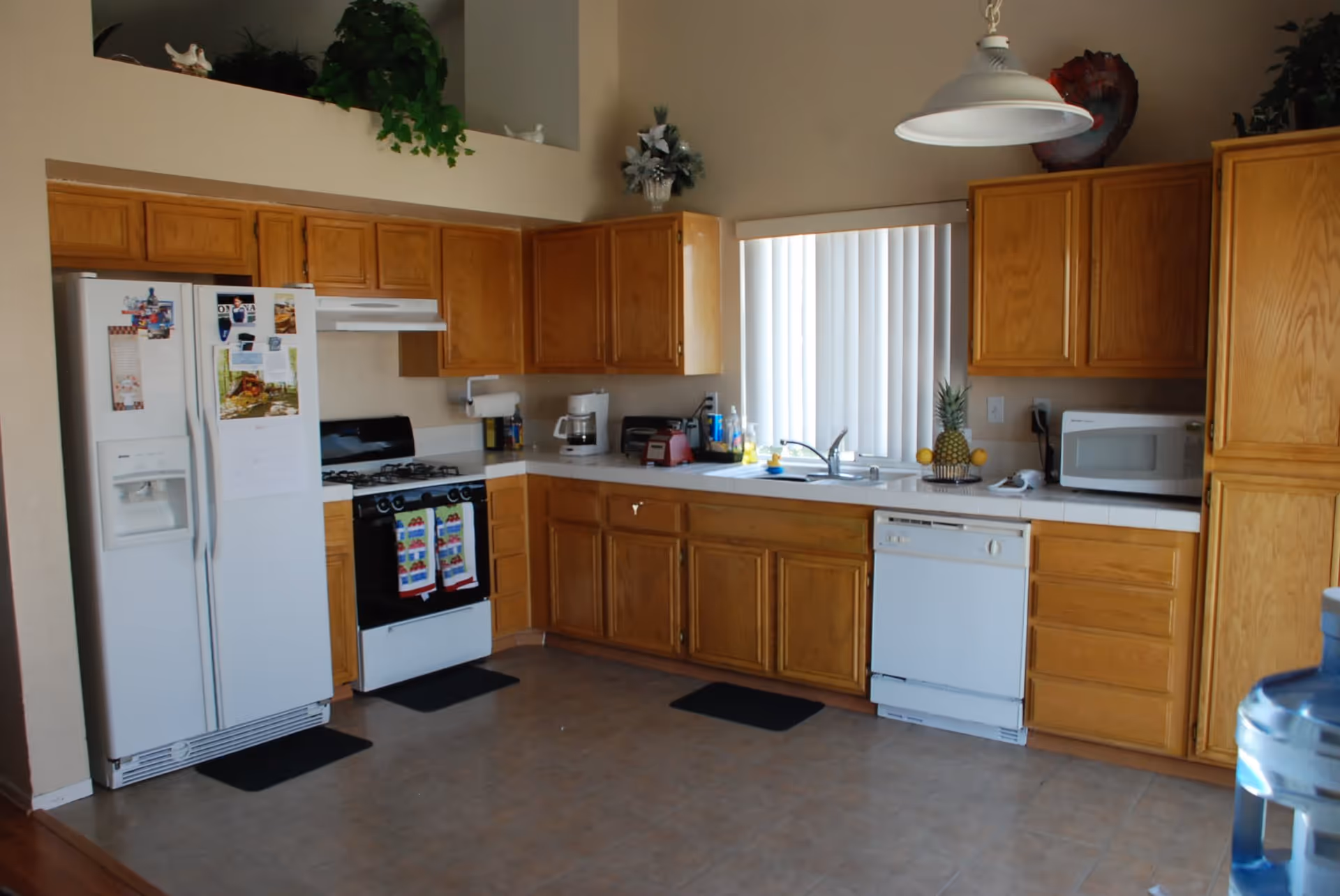 A kitchen with wooden cabinets, a white refrigerator with magnets and papers, a white gas stove with colorful towels hanging on the handle, a coffee maker, a toaster, a blender, a sink under a window with vertical blinds, a dishwasher, a microwave, and a water dispenser in the foreground.
