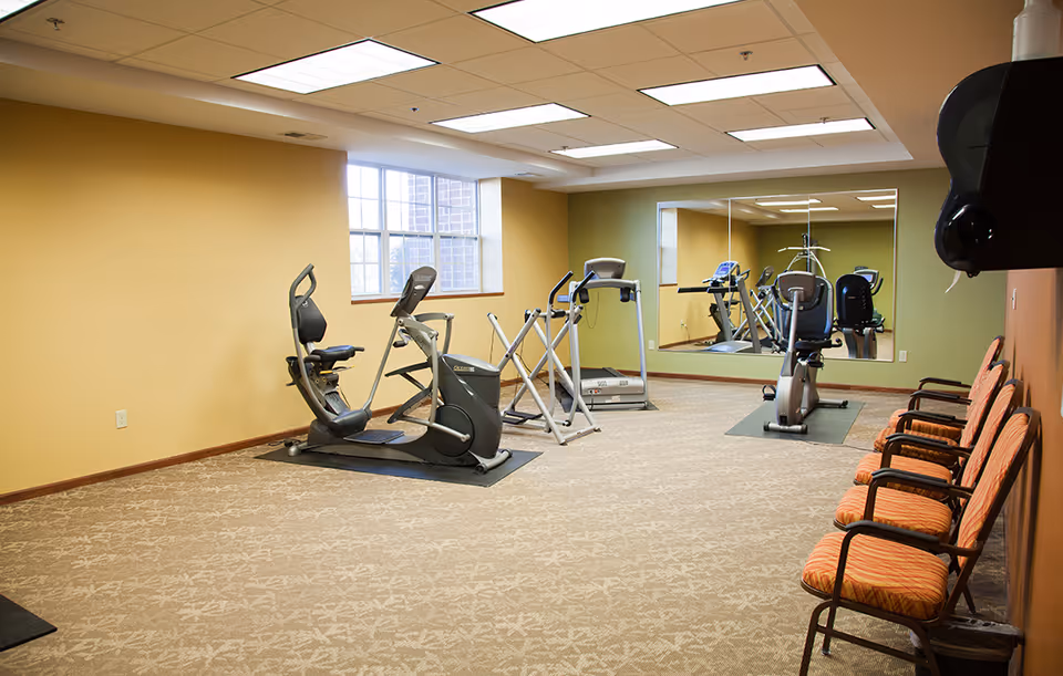 A small fitness room with exercise equipment including a recumbent bike, treadmill, and elliptical machine. There is a large mirror on one wall and a row of orange cushioned chairs along another wall. The room has beige carpet, light yellow and green walls, and a window letting in natural light.