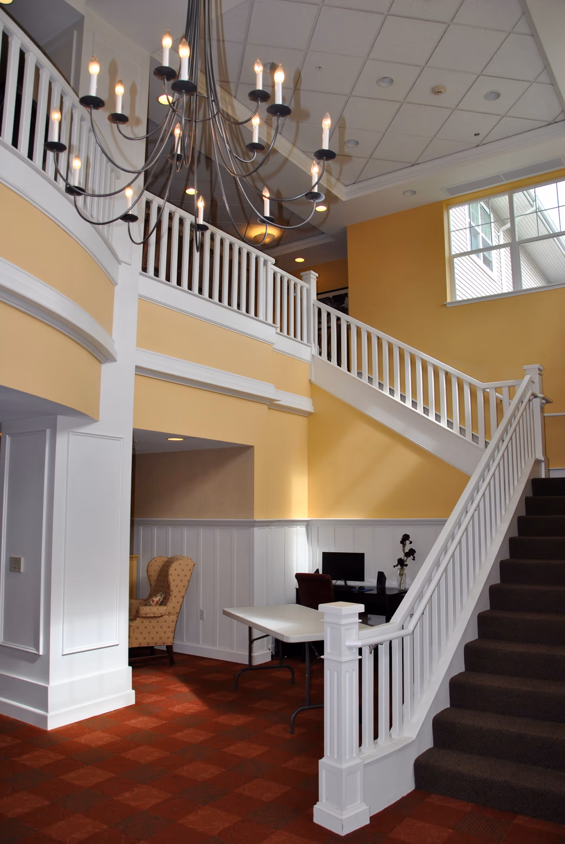 Interior view of a senior living facility featuring a staircase with white railings leading to an upper floor. The walls are painted yellow with white wainscoting, and there is a large window letting in natural light. A chandelier with multiple candle-like lights hangs from the ceiling. Below the staircase, there is a table, a computer desk with a monitor, and a patterned armchair in a cozy nook.
