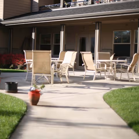 Outdoor patio area with several beige chairs and tables arranged on a concrete surface, adjacent to a building with multiple windows and a door. There is a curved walkway leading to the patio, with green grass and a small potted plant in the foreground.