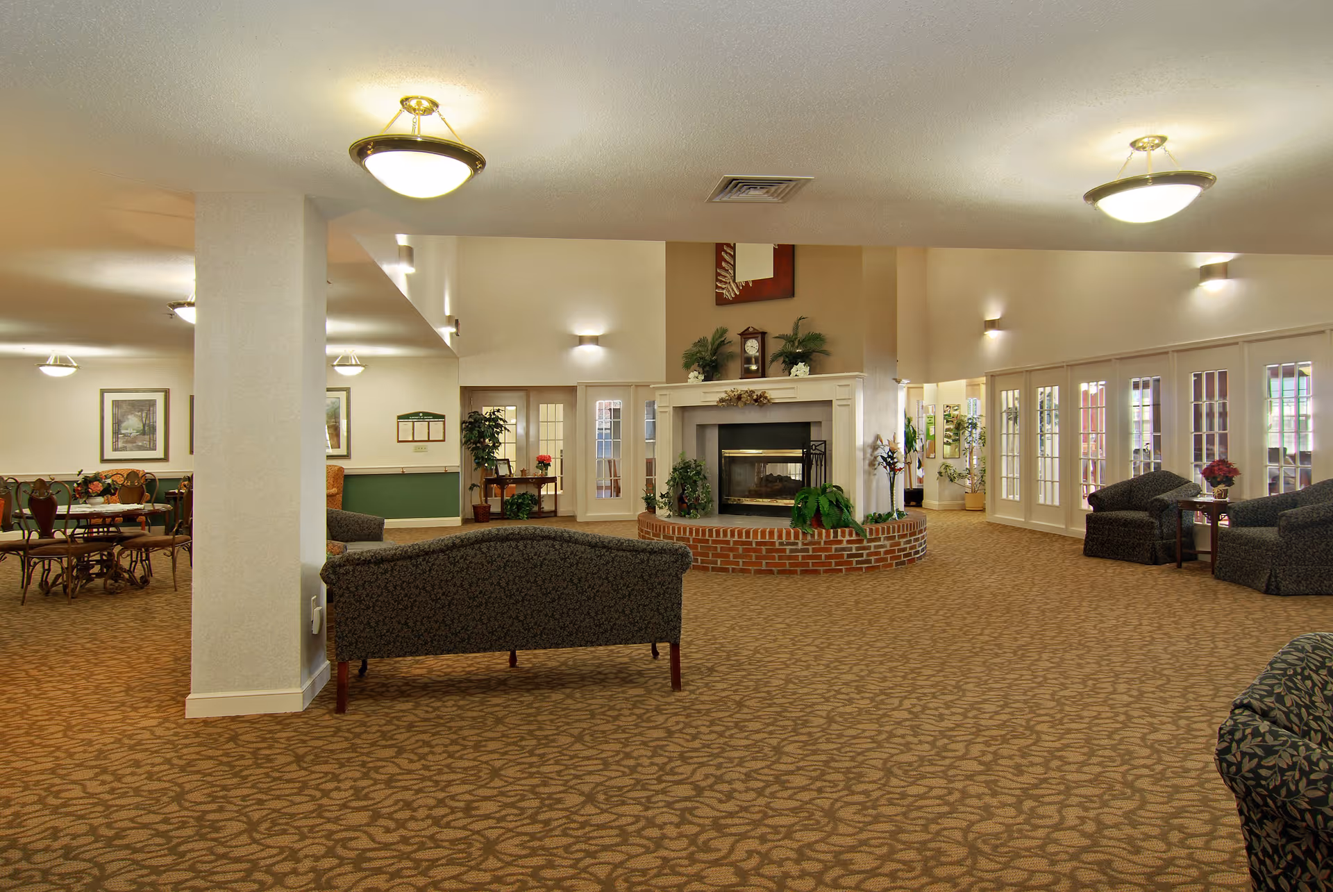 Spacious senior living facility common area with patterned carpet, multiple seating arrangements including sofas and chairs, a central fireplace with plants and decorative items, and large windows with glass doors allowing natural light.