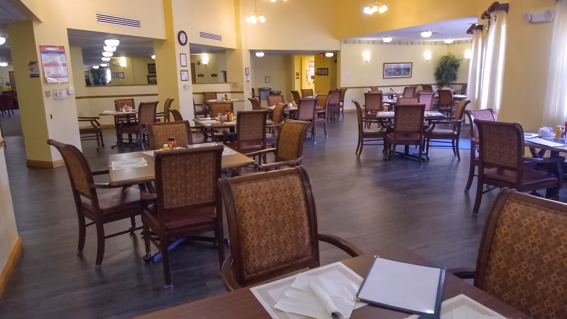 Spacious dining room with multiple tables and patterned chairs arranged across a large room with warm yellow walls and wood flooring.
