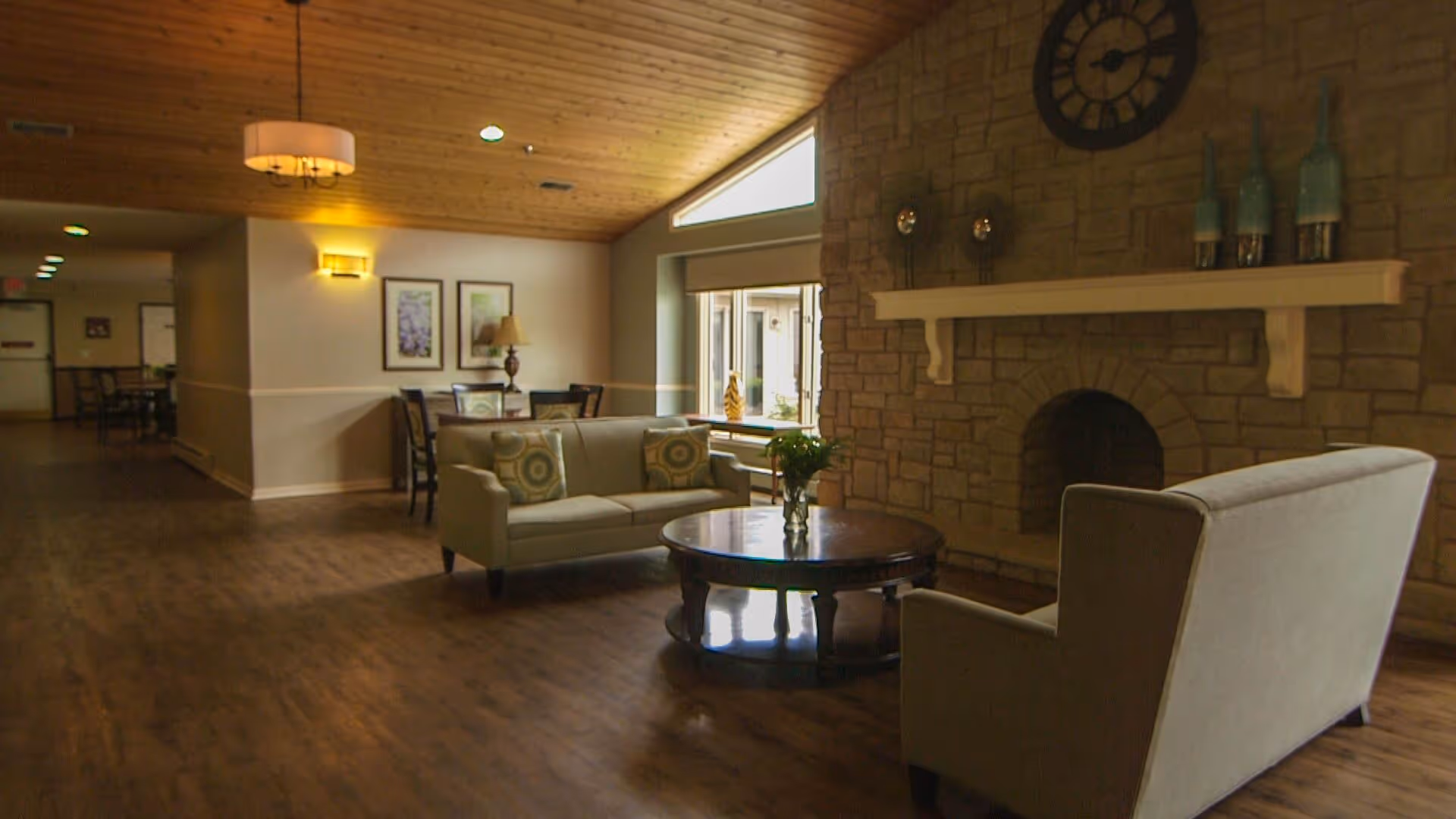 A cozy living room area with wooden flooring and a wooden ceiling. The room features a stone fireplace with a mantel decorated with blue vases and a large clock above it. There are two beige sofas with patterned cushions facing a round wooden coffee table with a vase of flowers. In the background, there is a dining area with a table, chairs, and framed artwork on the wall. The room is softly lit with wall sconces and a hanging light fixture.