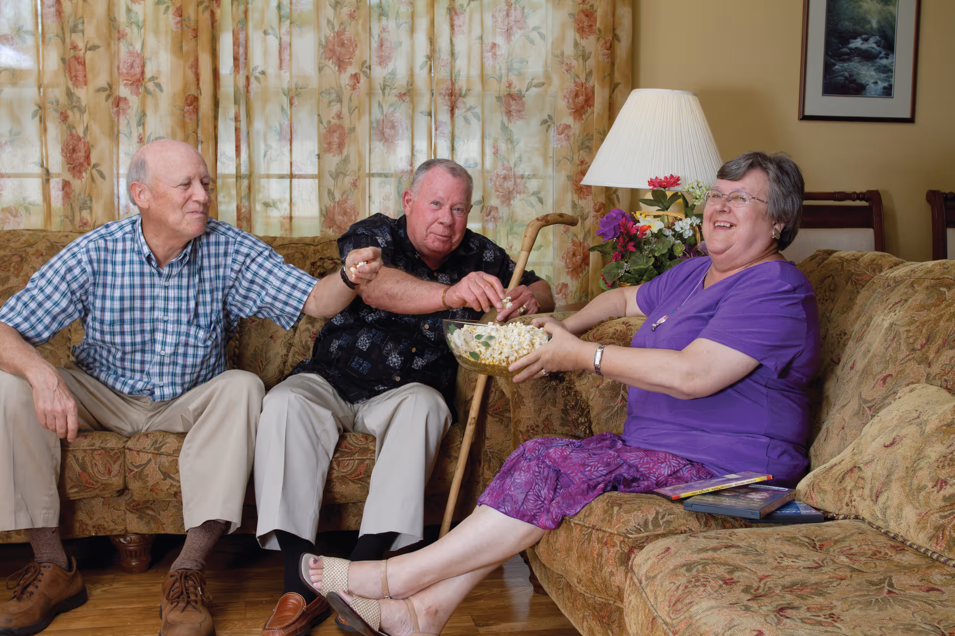 Three elderly people sitting on patterned sofas in a living room. Two men are sitting on one sofa, one wearing a checkered shirt and the other a dark patterned shirt holding a cane. A woman in a purple top and skirt is sitting on another sofa, holding a bowl of popcorn and smiling. The room has floral curtains, a table lamp, and a framed picture on the wall.