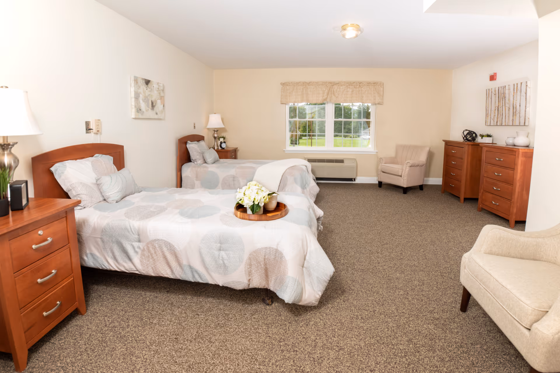 A bright and spacious senior living bedroom with two twin beds featuring light-colored patterned bedding. Each bed has a wooden headboard and is accompanied by a wooden nightstand with a lamp. The room has beige walls, a large window with a beige valance, two beige armchairs, and two wooden dressers. The carpet is a neutral brown tone, and there are decorative items and artwork on the walls and furniture.