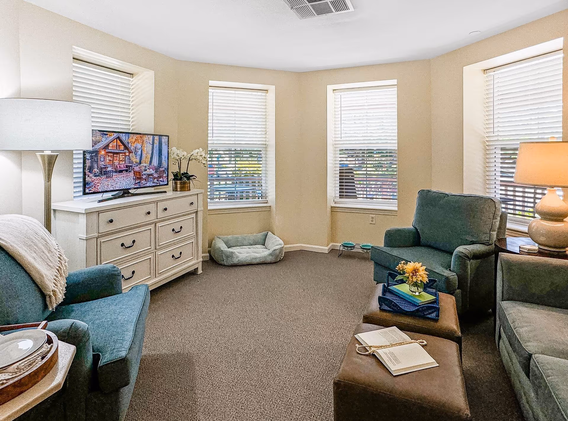 Bright sitting room with upholstered chairs, ottoman, TV on a dresser, and three windows with blinds.