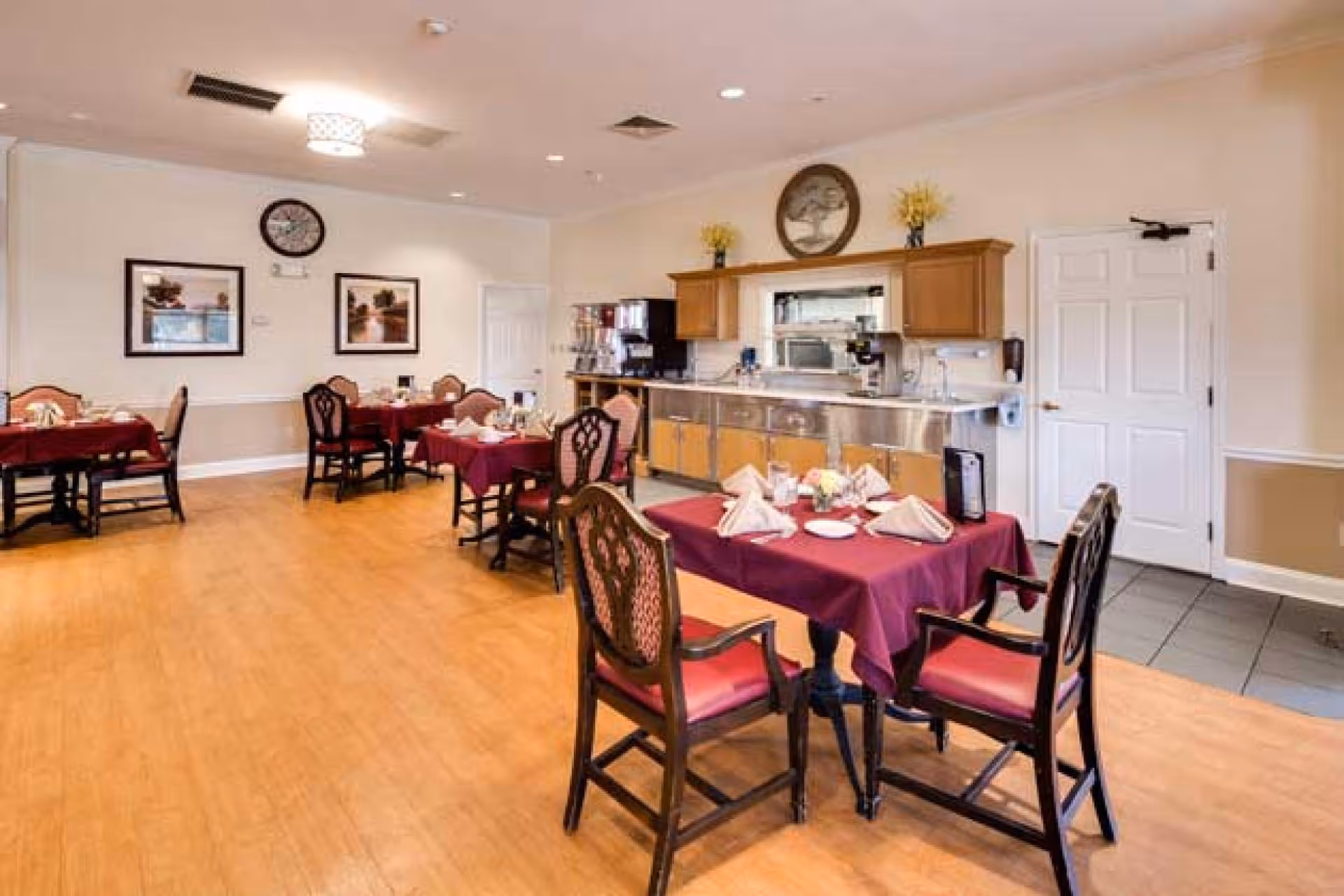 Dining room with set tables covered in burgundy tablecloths, wooden chairs, and a serving counter along the back wall.