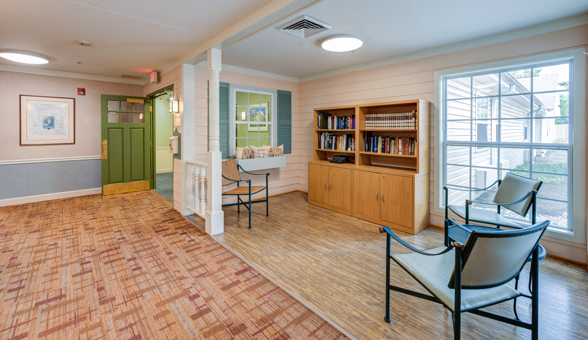 Well-lit lounge area in a memory care facility with chairs, a bookshelf, a large window, and a green entry door.