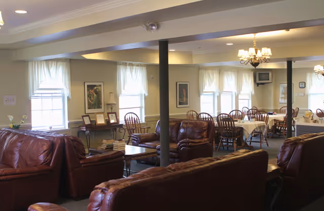 Interior view of a senior living facility common area with brown leather sofas and armchairs arranged around coffee tables. In the background, there are dining tables with chairs set near windows with white curtains. The room is softly lit by chandeliers and has framed artwork on the walls.