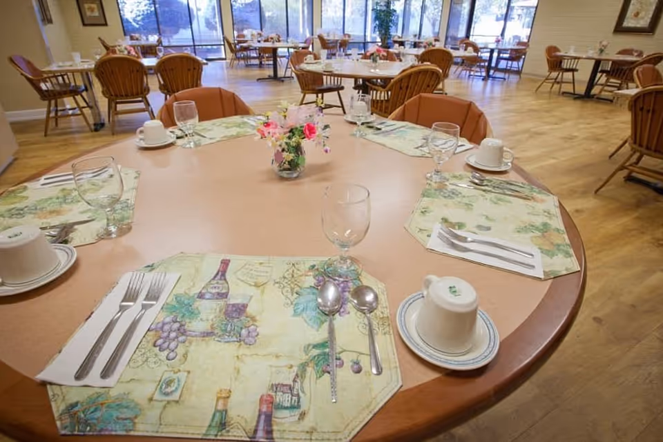 A dining room in a senior living facility with round tables set for meals. Each table has placemats, utensils, cups, glasses, and a small flower arrangement in the center. Wooden chairs surround the tables, and large windows in the background let in natural light.