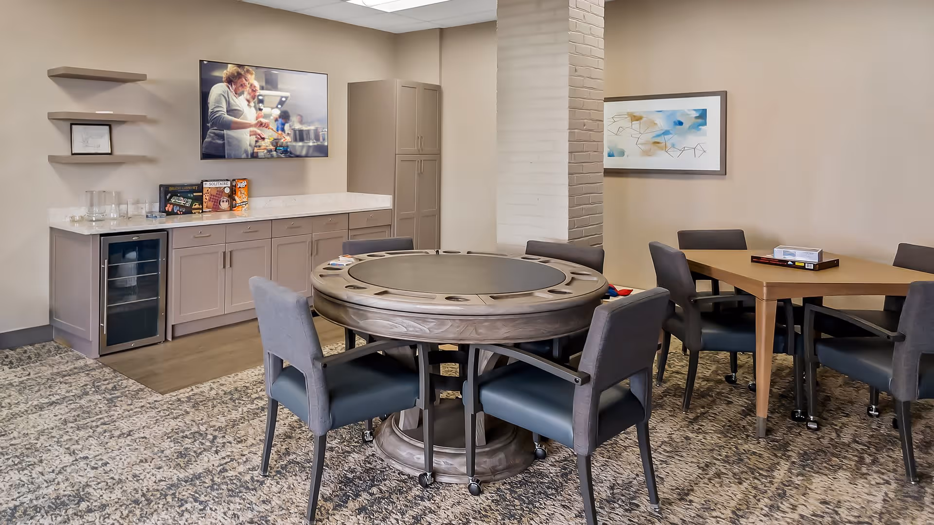 A well-lit common room with a round gaming table surrounded by six cushioned chairs on wheels. Behind the table is a countertop with cabinets, a small wine fridge, and board games on display. A flat-screen TV mounted on the wall shows an image of two people cooking. Another rectangular table with chairs is visible in the background, along with a framed abstract painting on the wall.