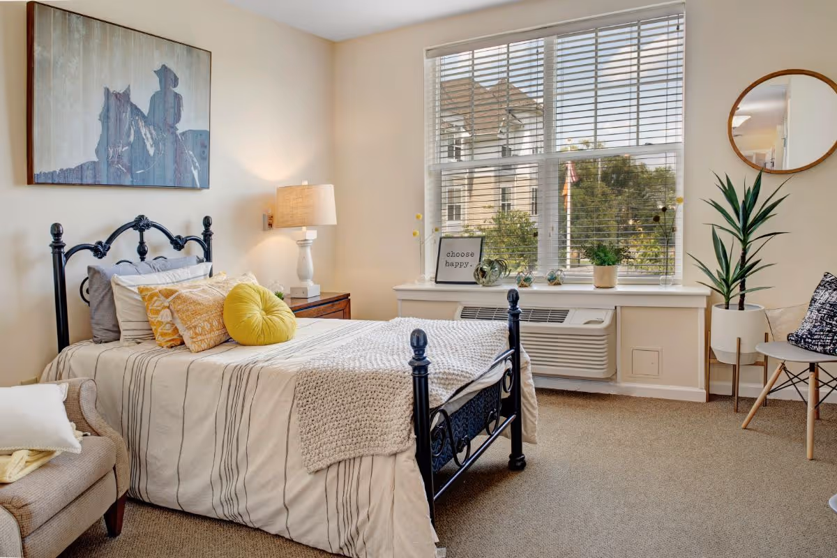 A cozy bedroom with a black metal bed frame, a bed made with striped bedding and multiple pillows including a round yellow one. There is a wooden nightstand with a lamp, a beige armchair with a pillow and blanket, a large window with blinds, and a windowsill decorated with plants and a framed sign that says 'choose happy.' A round mirror and a tall potted plant are on the right side of the room.