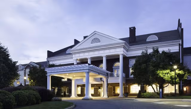 Front exterior view of a large, stately building with white columns and a covered entrance, surrounded by trees and bushes, illuminated at dusk.