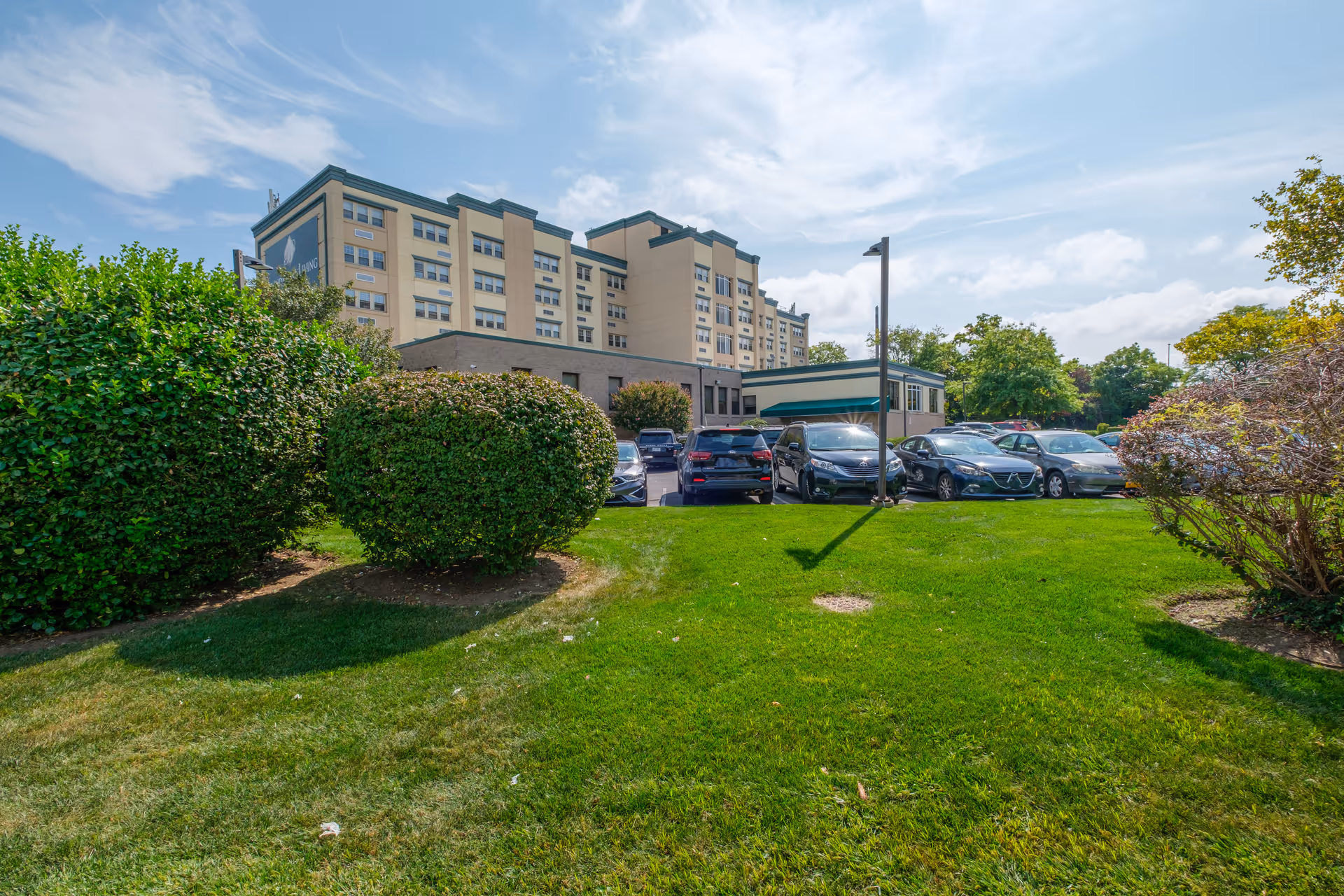 Front exterior of a multi-story senior living building with a grassy lawn, trimmed shrubs and a row of parked cars.