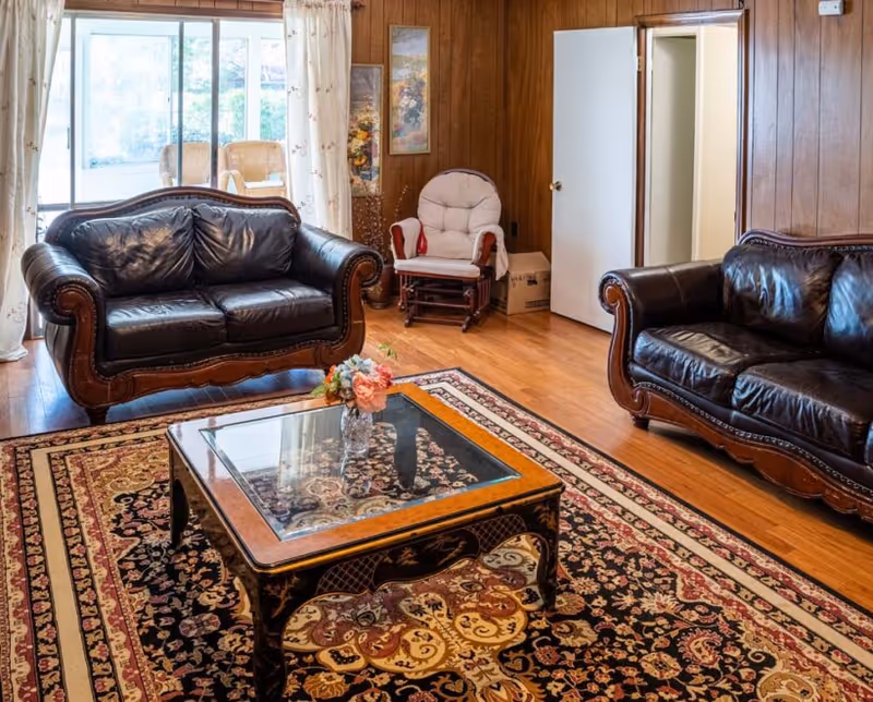 A cozy living room with two dark leather sofas facing each other, a glass-top wooden coffee table with a floral arrangement, a patterned area rug, wood-paneled walls, a cushioned rocking chair, and a sliding glass door with curtains leading to an outdoor area.