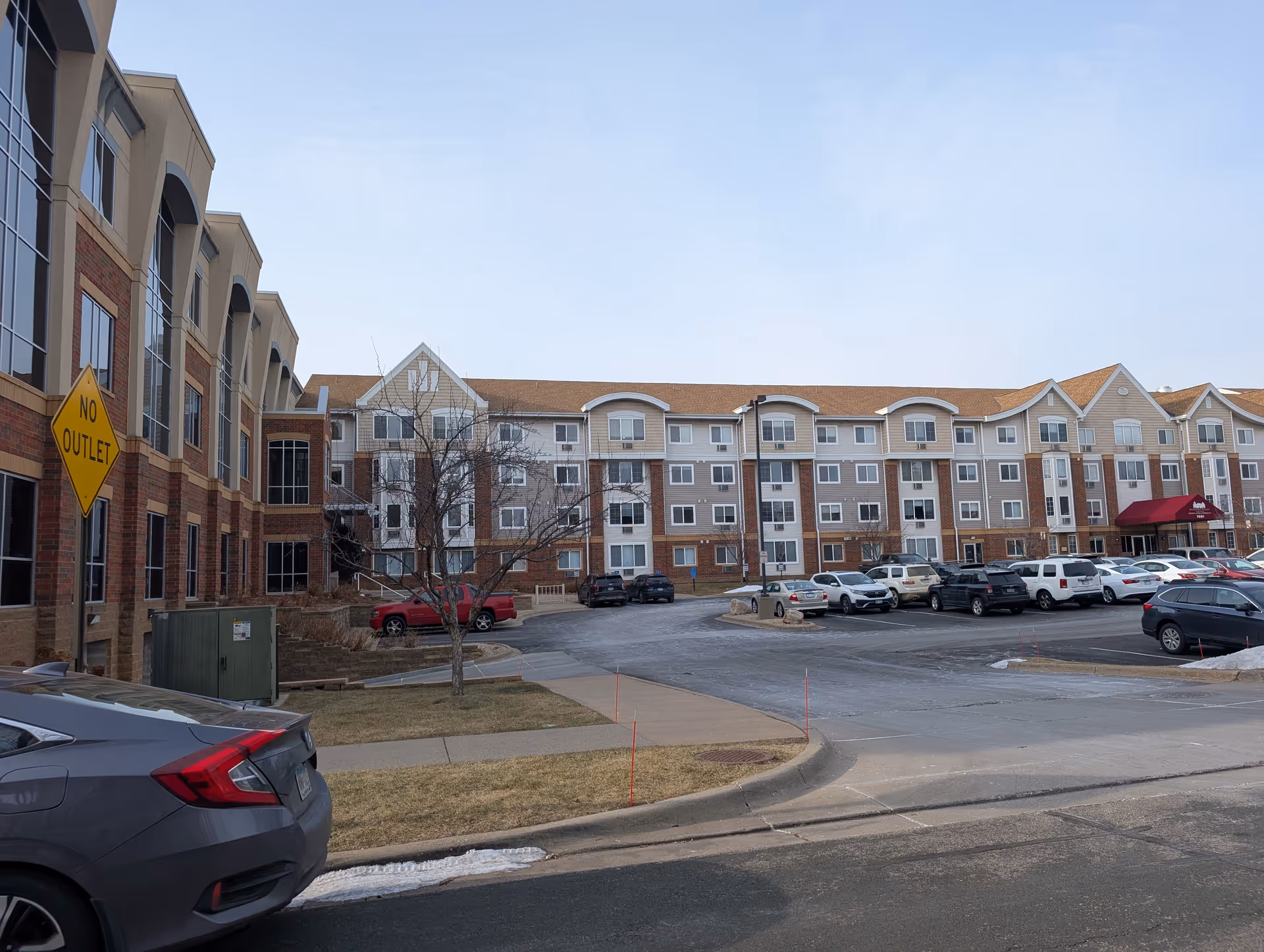 Front exterior of a multi-story senior living building with a parking lot, parked cars, and a 'No Outlet' sign.