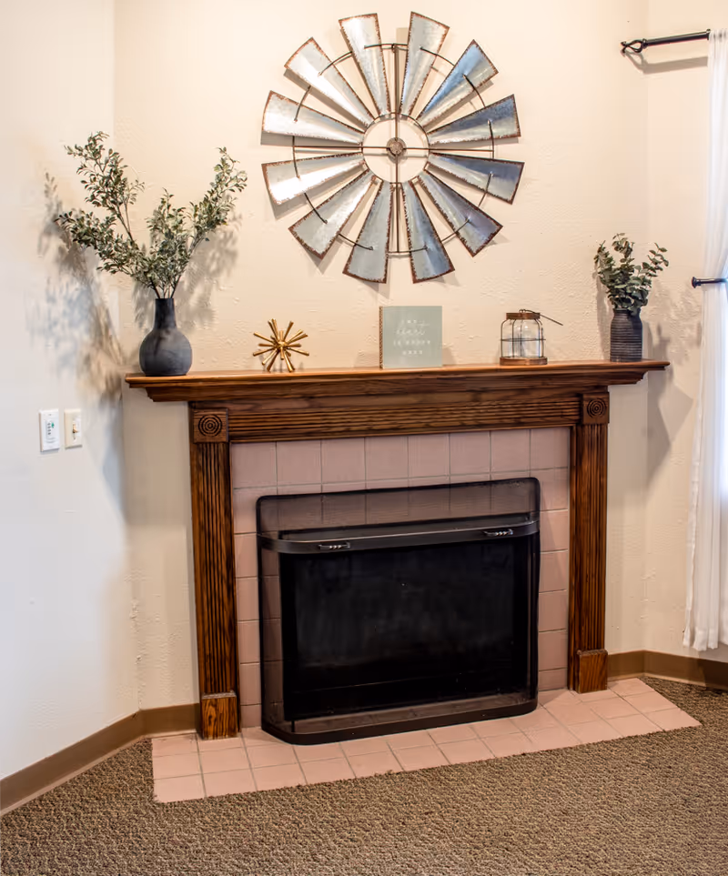 A cozy corner of a room featuring a wooden fireplace mantel with a black fireplace insert. The mantel is decorated with two vases containing green leafy branches, a small decorative golden star, a lantern, and a small sign. Above the mantel hangs a large metal wall decoration resembling a windmill. The floor is carpeted and there is a tiled hearth in front of the fireplace.