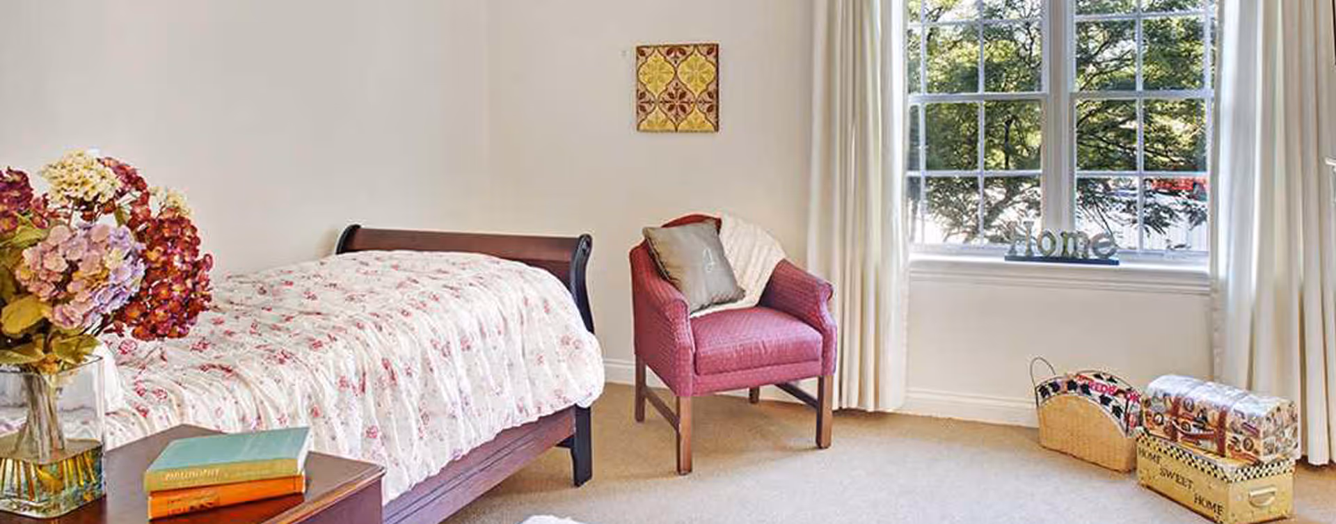 Sunlit bedroom with a floral-covered twin bed, a pink armchair, a bedside table with books and a vase of flowers, and a window ledge displaying a "Home" sign.