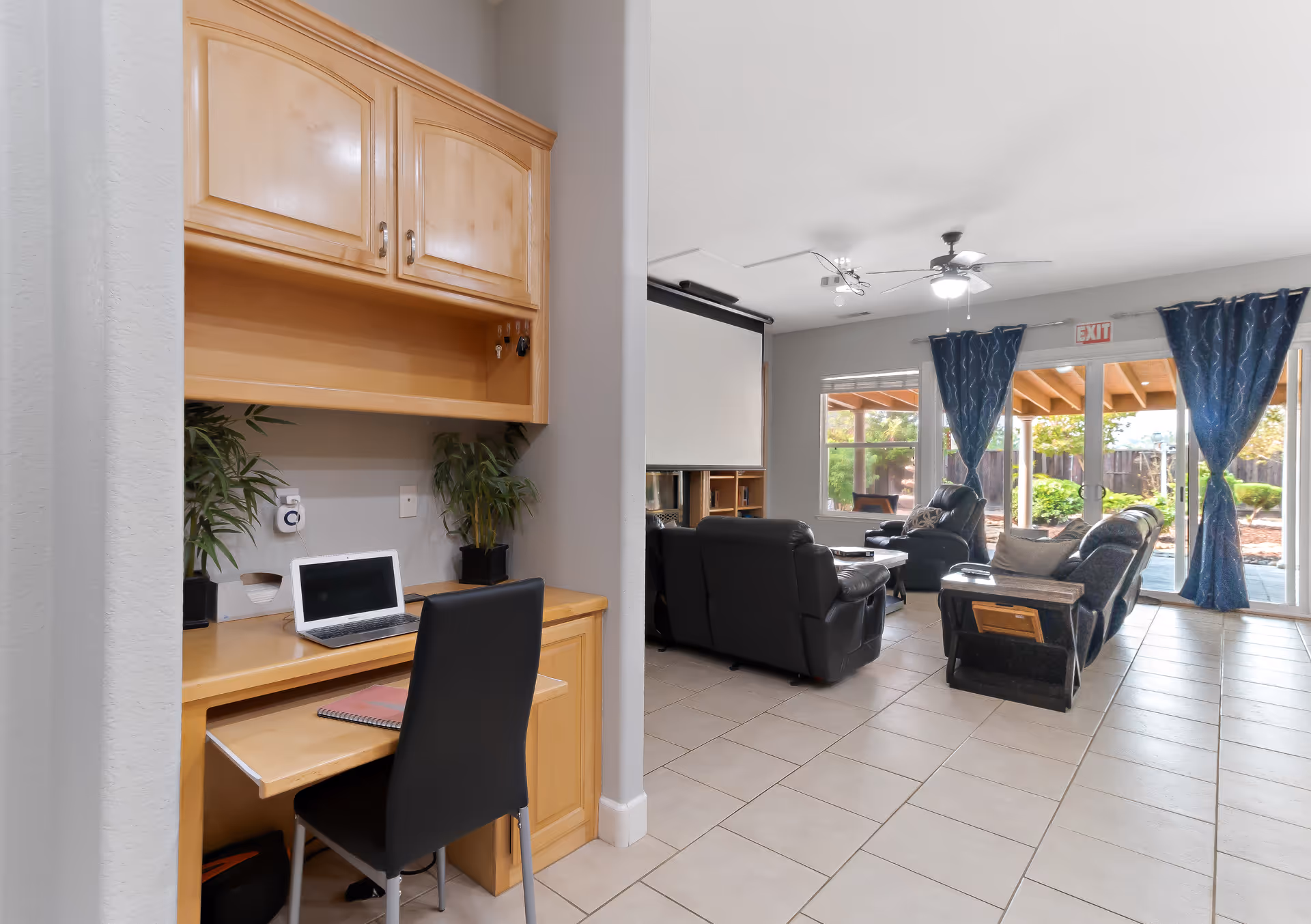 A bright living room with black leather sofas arranged around a coffee table, a ceiling fan with lights, large windows with blue curtains, and a sliding glass door leading to an outdoor patio. To the left, there is a small built-in wooden desk area with a laptop, a notebook, and two potted plants.