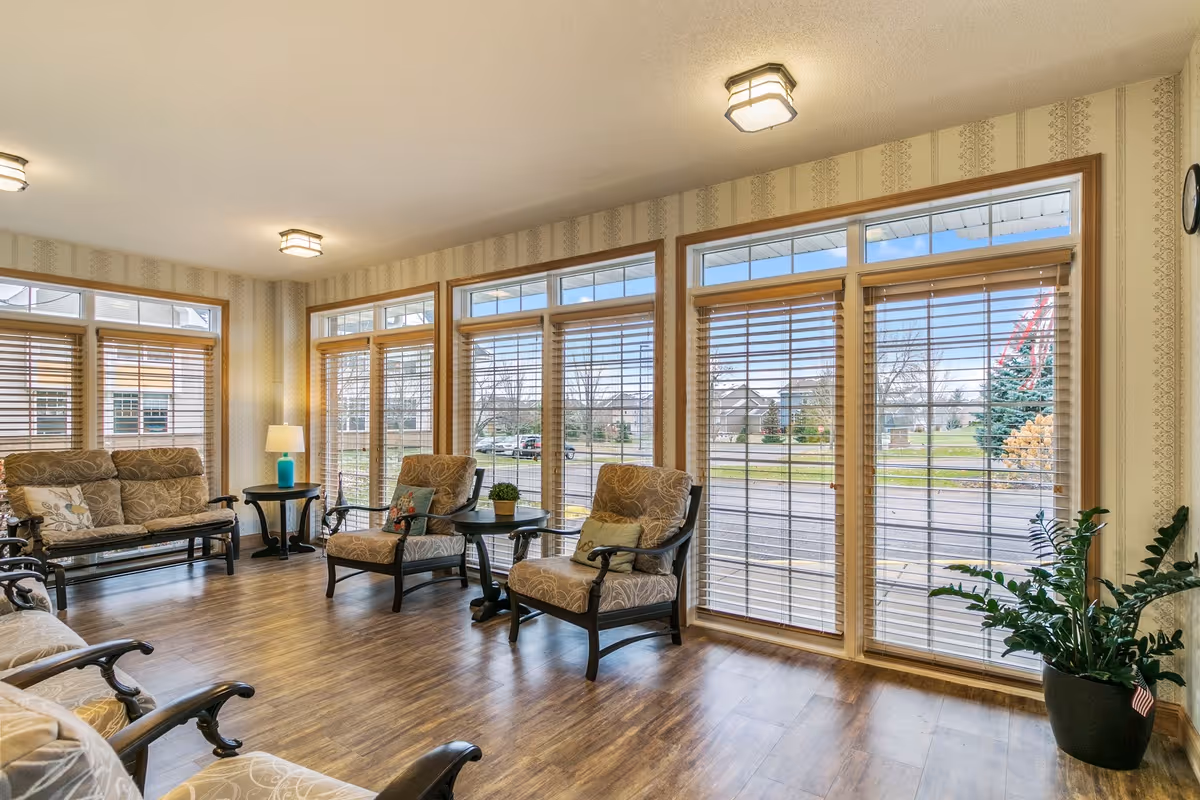 Bright lounge area with upholstered chairs and side tables arranged by large multi-pane windows with wood blinds overlooking the front lawn.