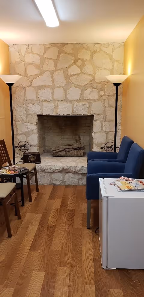 A cozy room with a stone fireplace in the center, flanked by two tall floor lamps with white shades. To the left, there are two wooden chairs with beige cushions and a small round table with magazines on it. To the right, there is a blue upholstered armchair next to a small white refrigerator with more magazines on top. The floor is wooden with a warm tone.