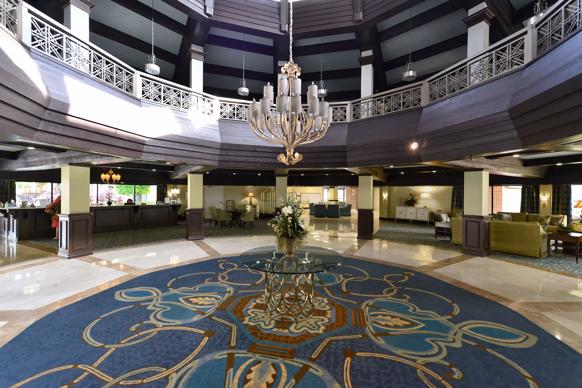 Spacious and elegant lobby area with a large decorative chandelier hanging from a high ceiling. The floor features a large blue and gold patterned rug with a glass table and floral arrangement in the center. Surrounding the lobby are seating areas with sofas and chairs, and a reception desk is visible in the background.