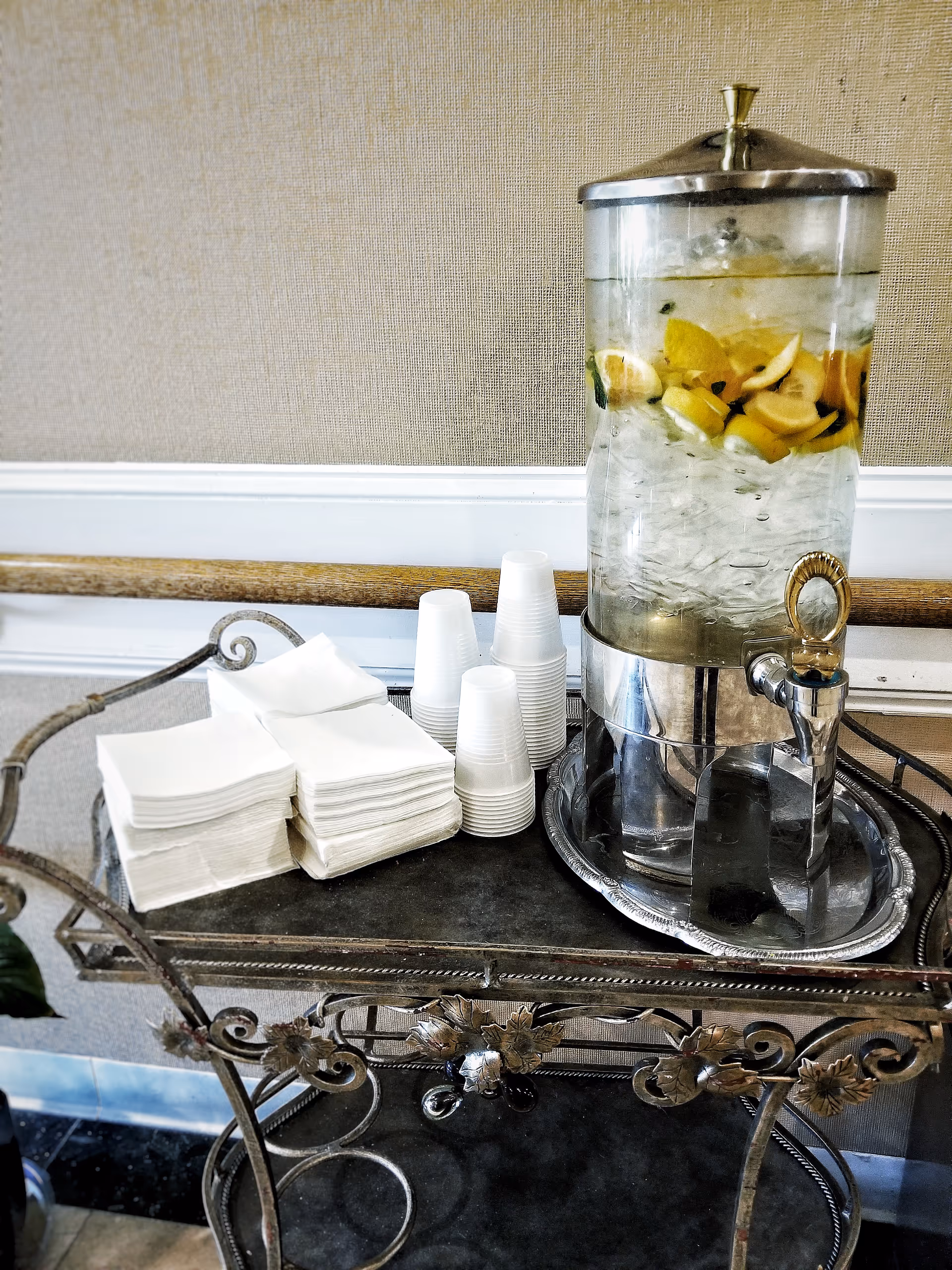 A beverage dispenser filled with ice water and lemon slices on a decorative metal cart. Next to the dispenser are stacks of white napkins and several white disposable cups. The background shows a beige textured wall with white trim.