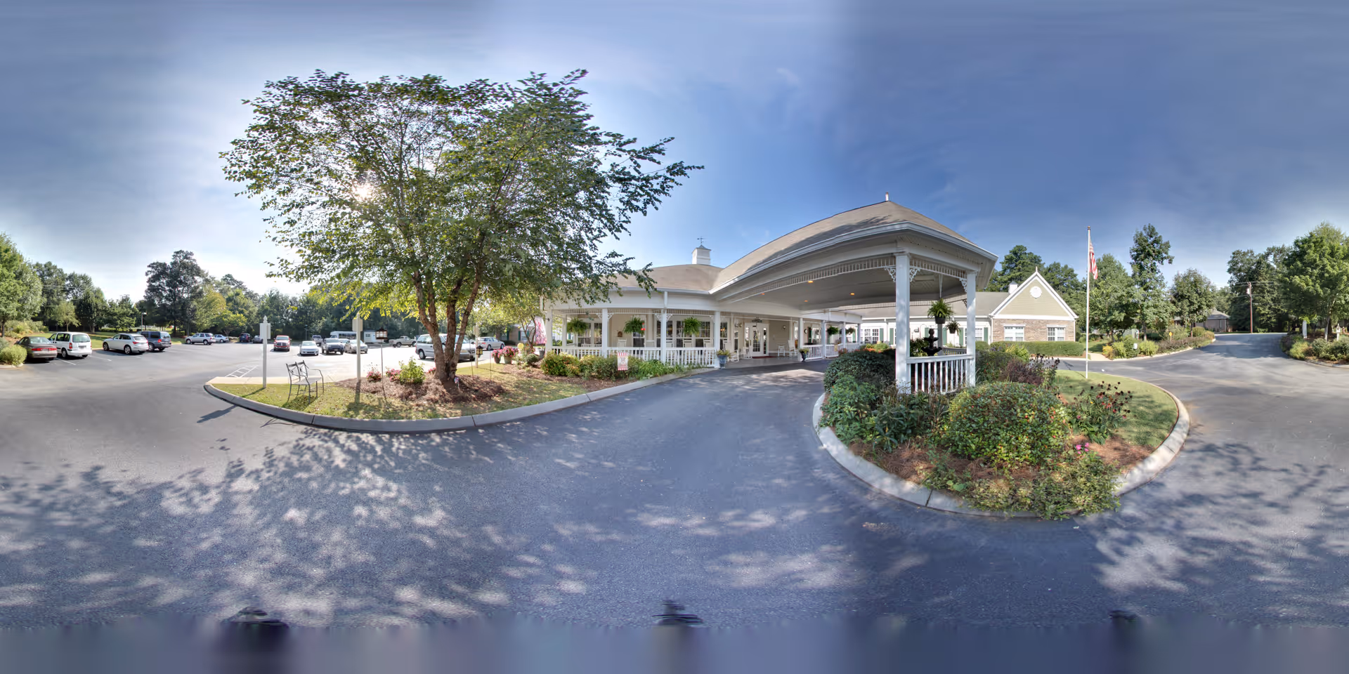 Front entrance of a senior living facility with a covered porte-cochère, circular driveway, parking lot, and landscaped trees and shrubs.