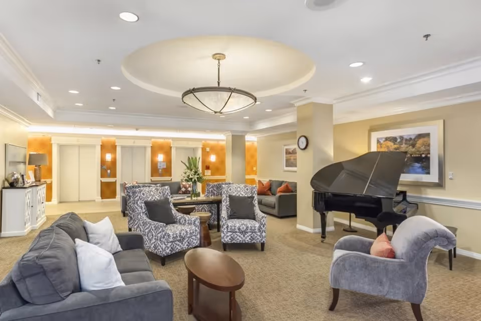 Well-lit seating area in a senior living facility lobby with sofas, patterned armchairs, a grand piano, and elevators in the background.
