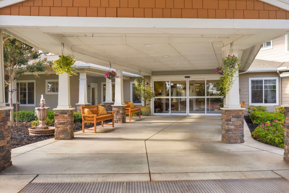 Covered entrance area of The Commons at Union Ranch facility with stone pillars, hanging flower baskets, wooden benches with cushions, a small fountain on the left, and automatic glass doors leading inside.