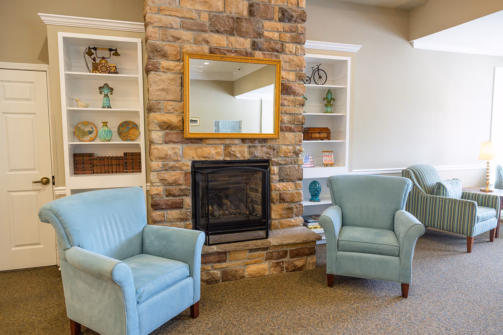 A comfortable common room with two light-blue armchairs in front of a stone fireplace flanked by white built-in shelves and additional seating.