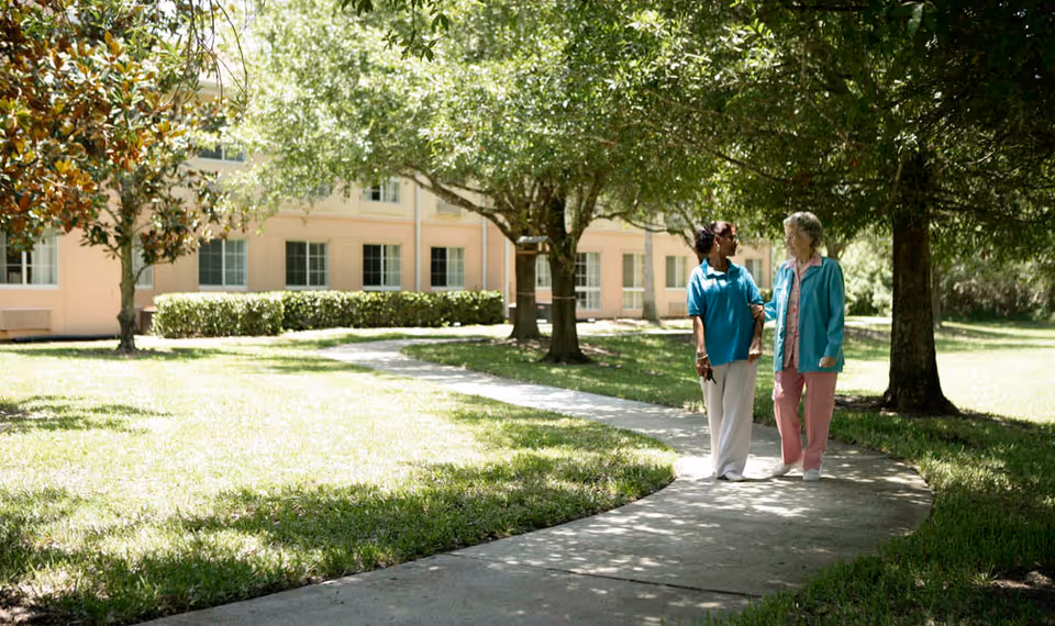 Two women walking arm in arm on a curved concrete path in a sunny garden area with green grass and trees, with a light-colored building with multiple windows in the background.
