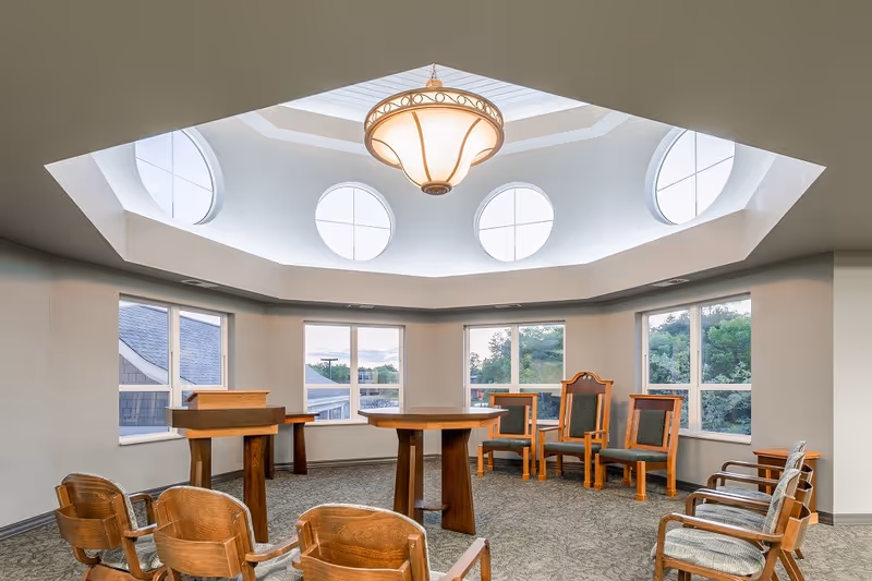 A bright, octagonal room with large windows and circular skylights. The room contains wooden chairs arranged in a semi-circle around a central wooden table and a wooden podium. The ceiling features a decorative hanging light fixture.