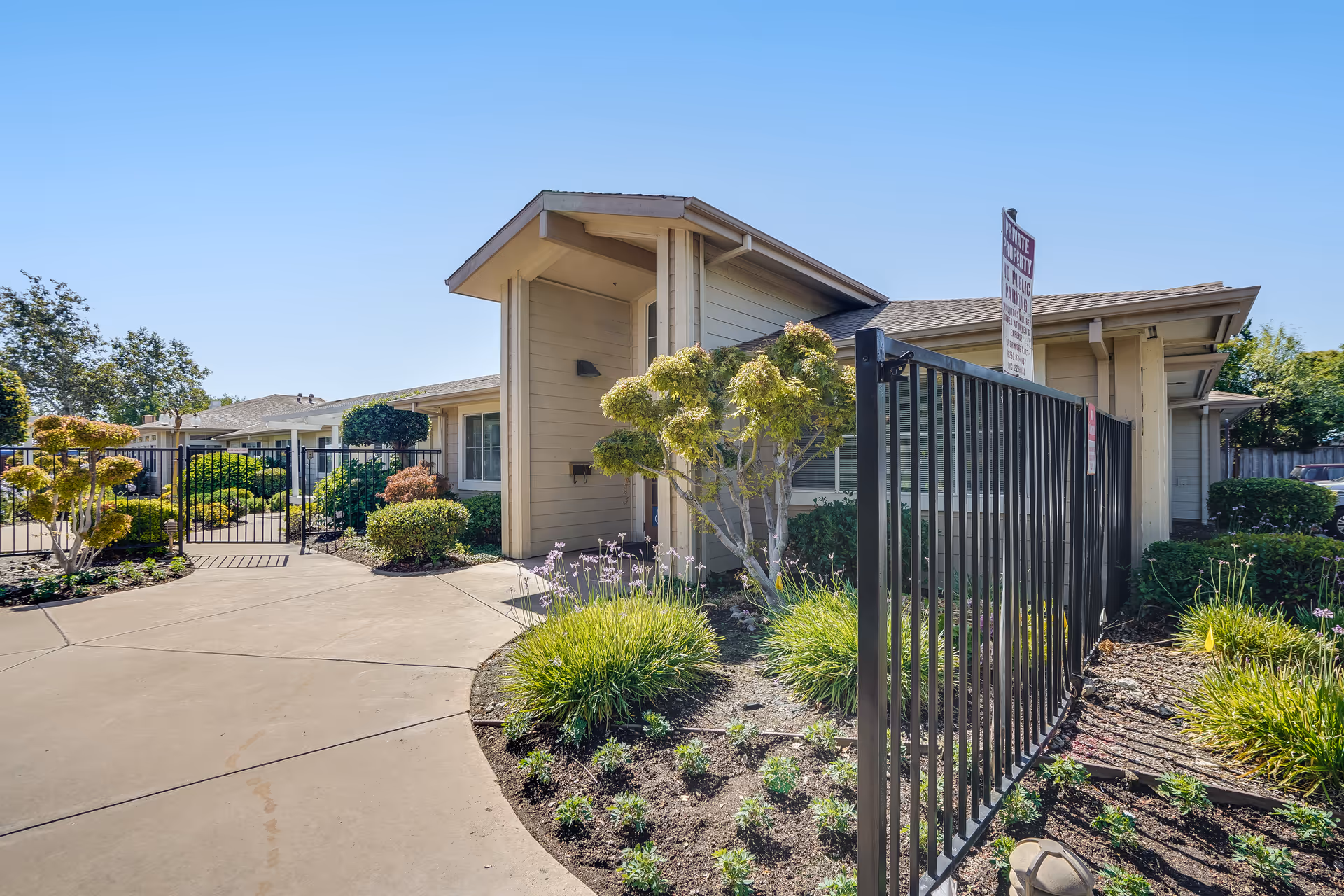 Exterior view of a single-story beige senior living building with a gated entrance, landscaped walkway, and shrubs under a clear blue sky.