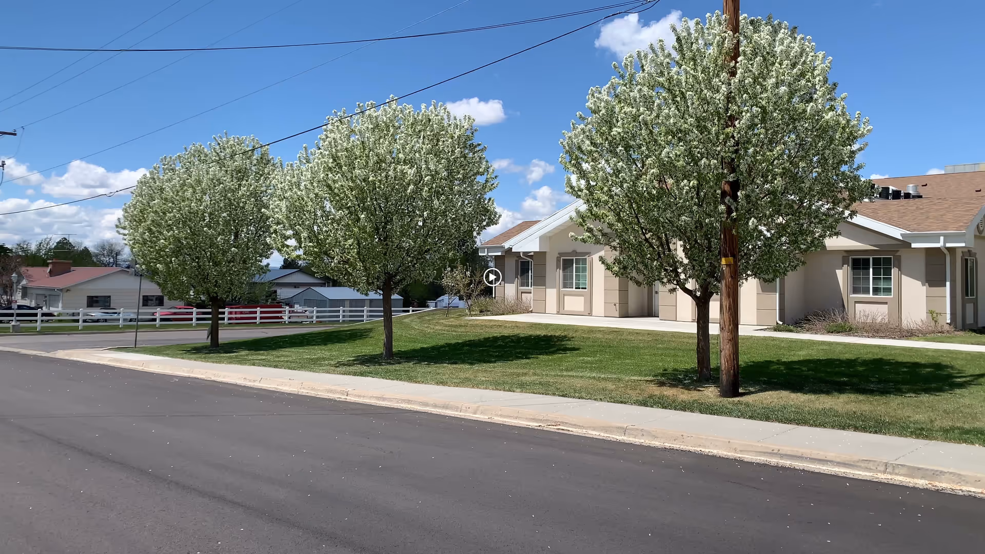 Exterior view of Cache Valley Assisted Living and Memory Care facility showing a single-story beige building with a brown roof, three blossoming trees with white flowers on a green lawn, a sidewalk, and a paved street under a blue sky with scattered clouds.