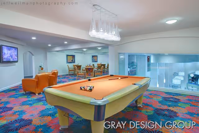 Interior recreational room with a pool table in the foreground, two orange armchairs to the left, and several tables and chairs in the background. The room has colorful floral carpet and white walls with framed artwork. A flat-screen TV is mounted on the left wall.
