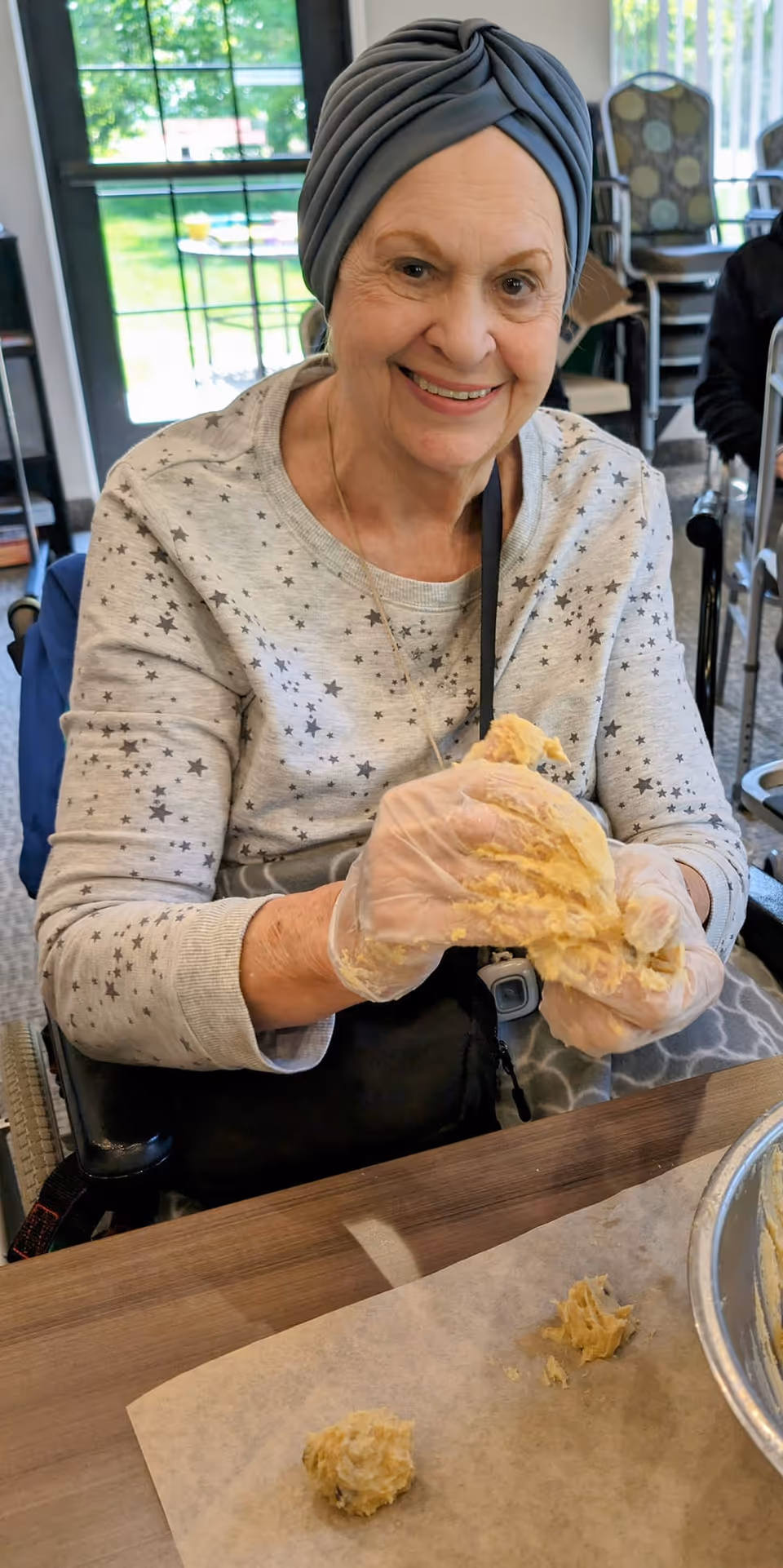 An elderly woman wearing a gray turban and a light gray sweater with star patterns is smiling while holding dough with gloved hands. She is seated at a table with parchment paper and dough balls in front of her, in a bright room with large windows and chairs in the background.