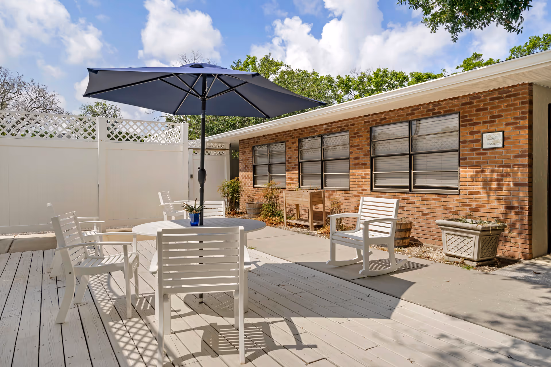 Outdoor patio area with white wooden chairs and a round table under a large blue umbrella. The patio is adjacent to a brick building with several windows and some potted plants along the wall. The sky is partly cloudy with trees visible in the background.