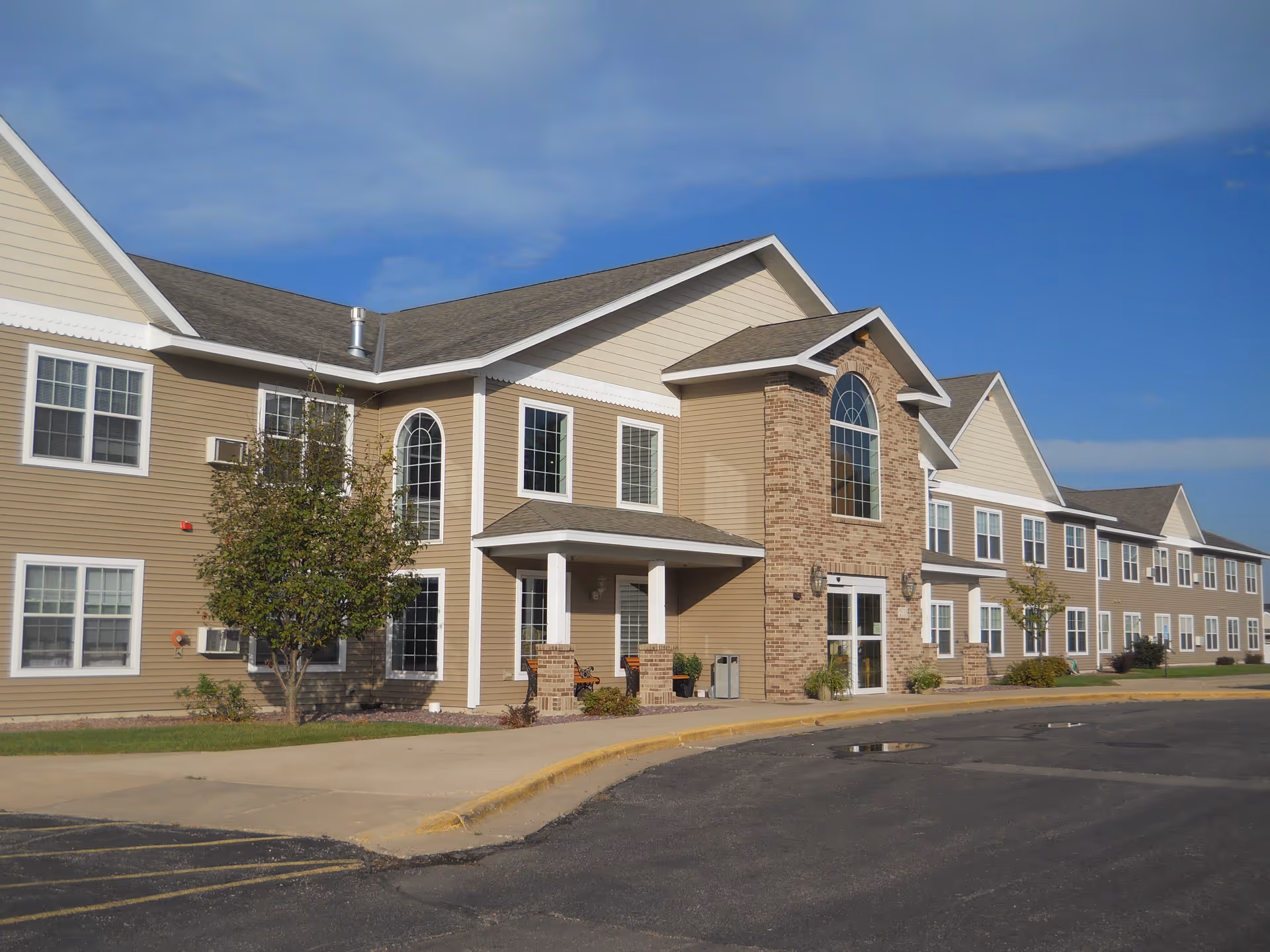 Two-story beige senior living building with large arched windows and a covered entrance under a clear blue sky.