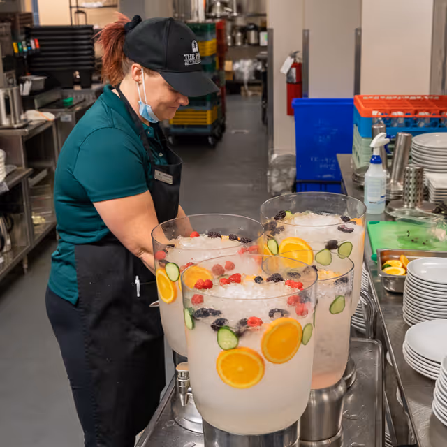 A staff member wearing a green shirt, black apron, and black cap with The Pines logo is preparing large beverage dispensers filled with ice, slices of orange, cucumber, and assorted berries in a kitchen setting.