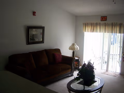 Living room with a brown couch, oval glass-top coffee table holding a potted plant, side table with a lamp, and a sliding glass door with vertical blinds and an EXIT sign.