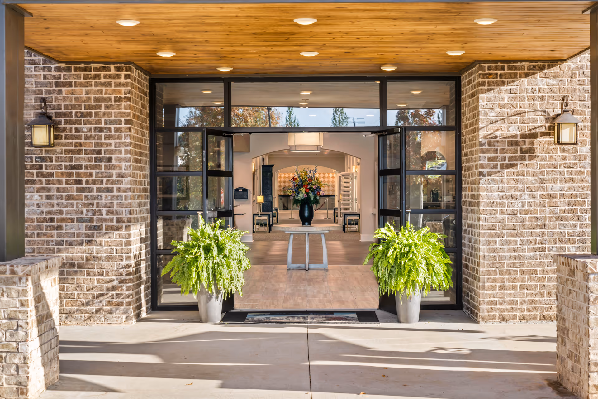 Entrance to a senior living facility with double glass doors framed by brick walls and two large potted ferns on either side. Inside, a table with a large floral arrangement is visible, along with a well-lit interior space featuring wooden flooring and modern decor.
