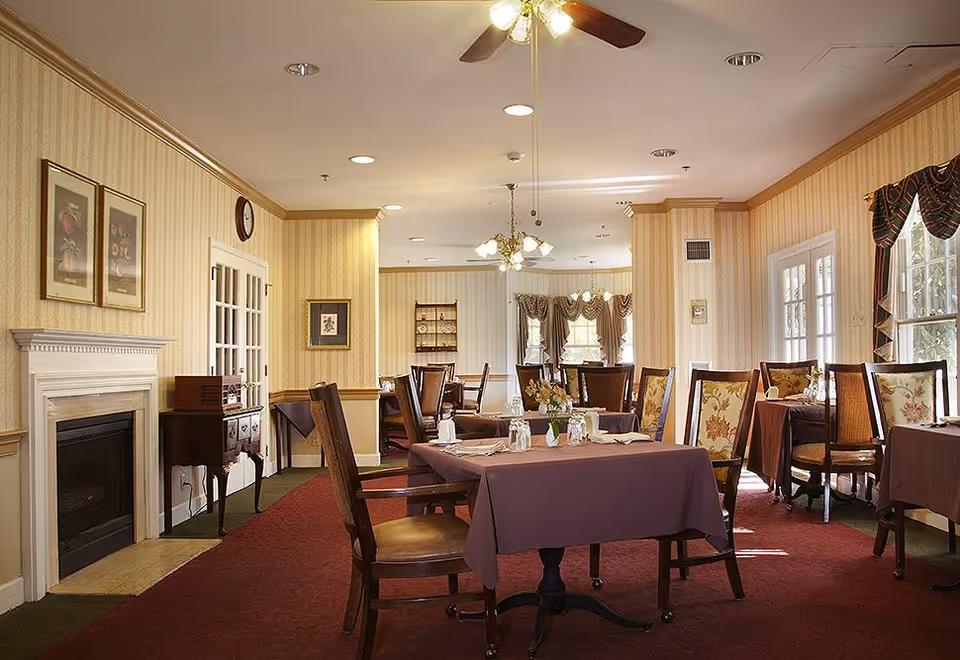 A dining room with several tables covered in purple tablecloths, each set with napkins, glasses, and small flower arrangements. The room features patterned wallpaper, a fireplace on the left wall, framed pictures, and large windows with draped curtains allowing natural light to enter. Ceiling fans and light fixtures hang from the ceiling.